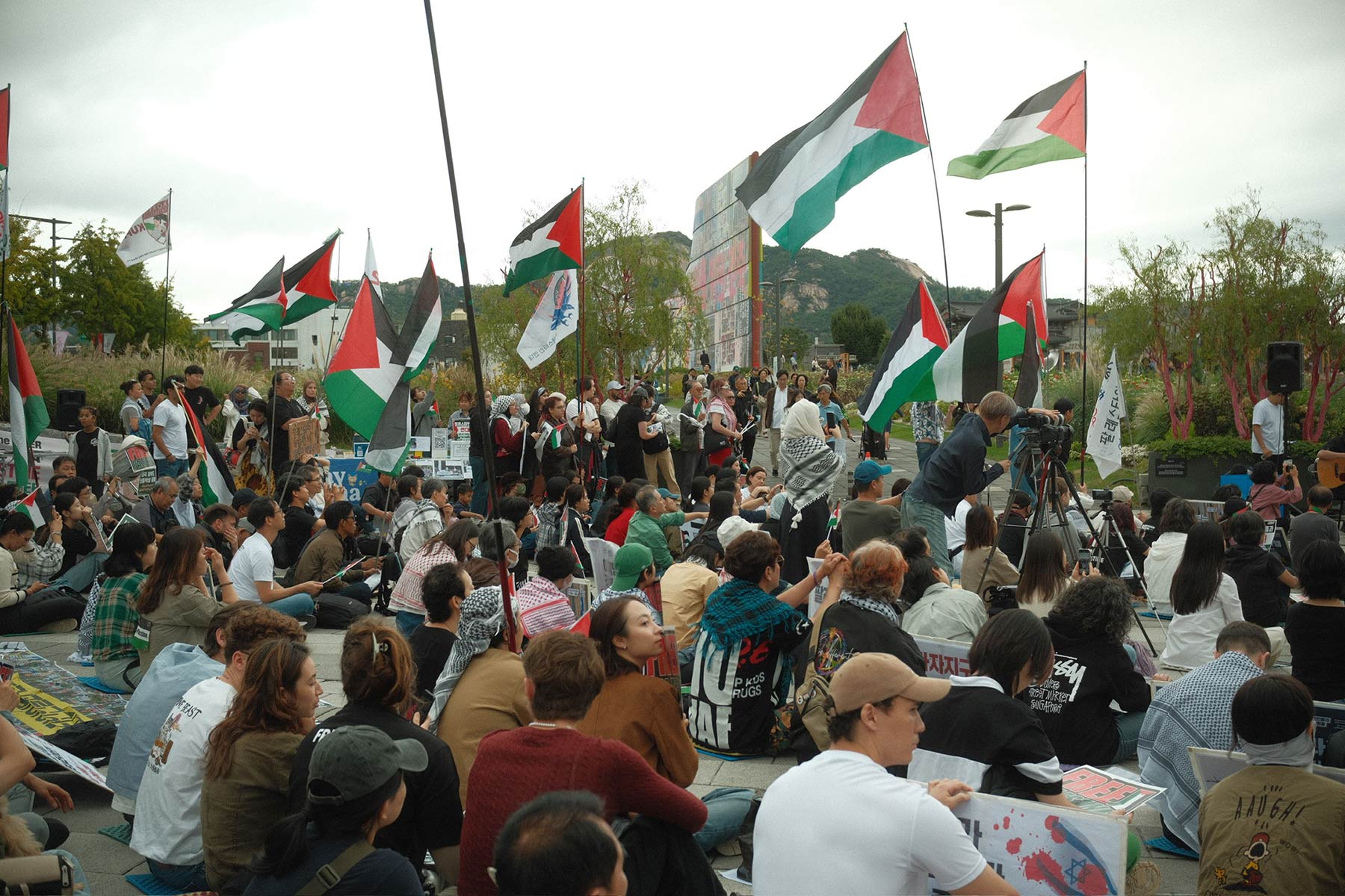 Hundreds of people sitting and standing peacefully at a protest with lots of Palestinian flags