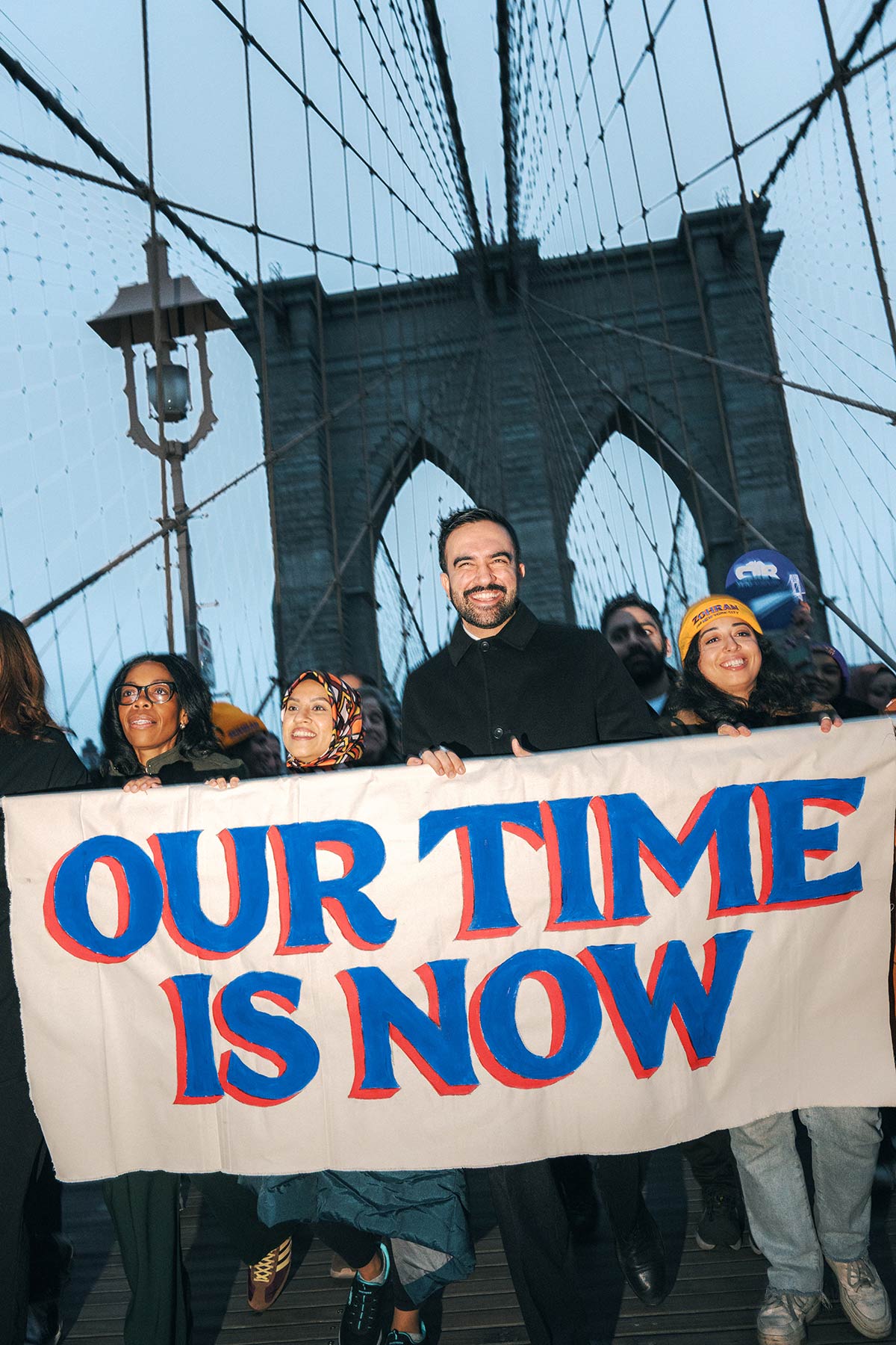 Zohran Mamdani leading a parade across the Brooklyn Bridge, holding a banner that reads "Our Time is Now"
