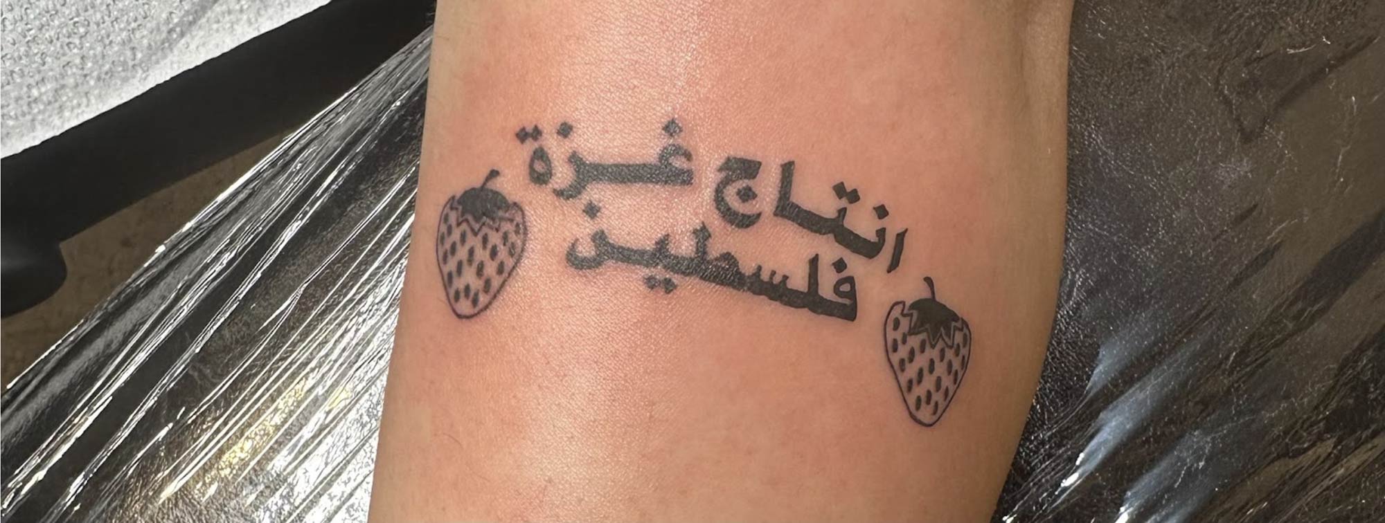 close-up of a tattoo in black on olive skin, a line-drawing strawberry on either side of arabic writing, ابتاج غزة فلسطين