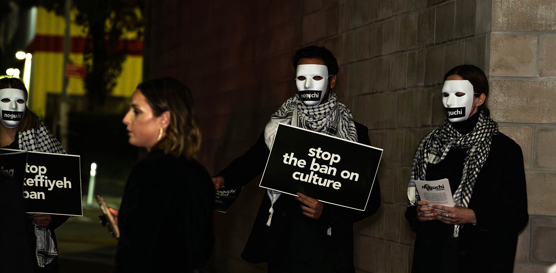 Night photo of Noguchi museum staff in black with white keffiyehs and white face masks, holding protest signs outside the annual Noguchi museum gala: "Stop the ban on culture","Stop the keffiyeh ban"